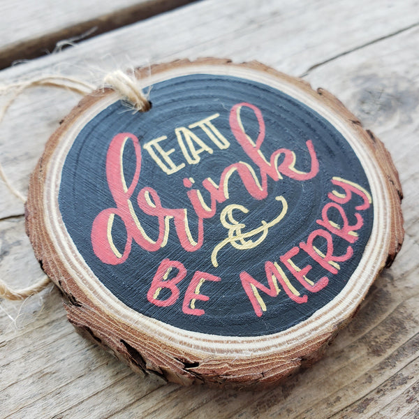 rustic wood slice ornament that says eat drink & be merry in gold and red hand lettering