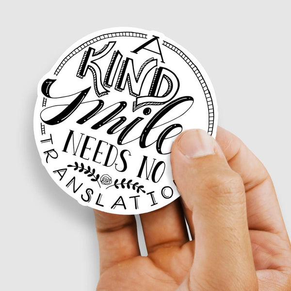 3" hand lettered illustrated vinyl sticker saying a kind smile needs no translation in black and white shown with a woman's hand holding the sticker