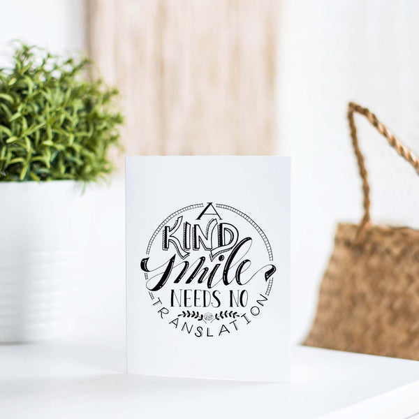 A Kind Smile Needs No Translation A2 Greeting Card black design on white card with white envelope shown on a table with a plant and handbag