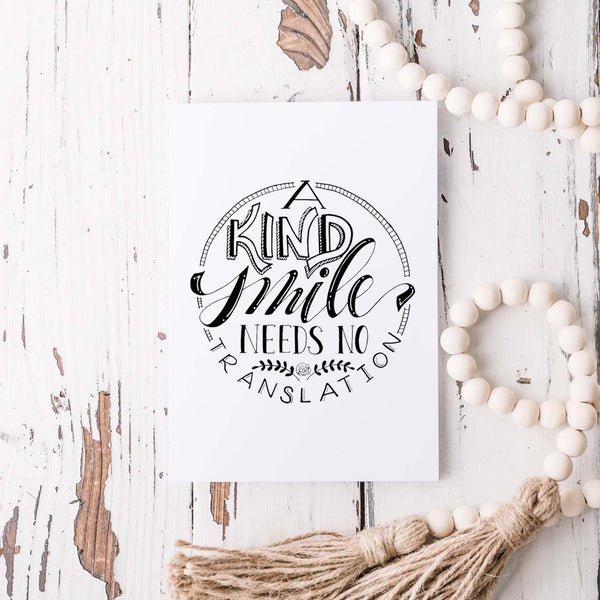 A Kind Smile Needs No Translation A2 Greeting Card black design on white card with white envelope shown on a rustic white wooden table with a wooden bead garland