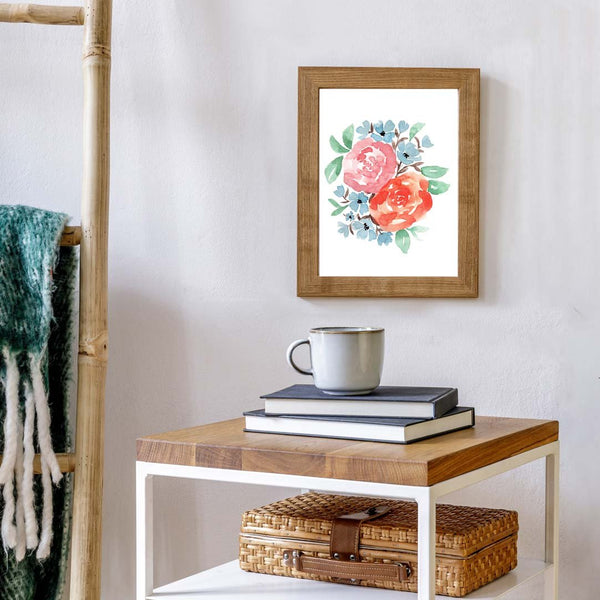 Watercolor wall art print with coral and rust colored roses, little muted blue accent flowers and green leaves shown hanging on a grey wall in a wooden frame above a table with a stack of books and coffee mug