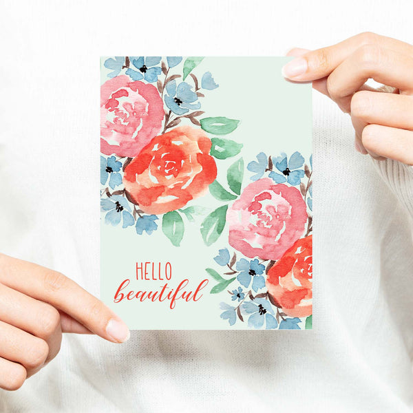 hello beautiful floral watercolor greeting card with coral, pink, blue flowers and green leaves with white A2 envelope shown with a woman in a white sweater holding card