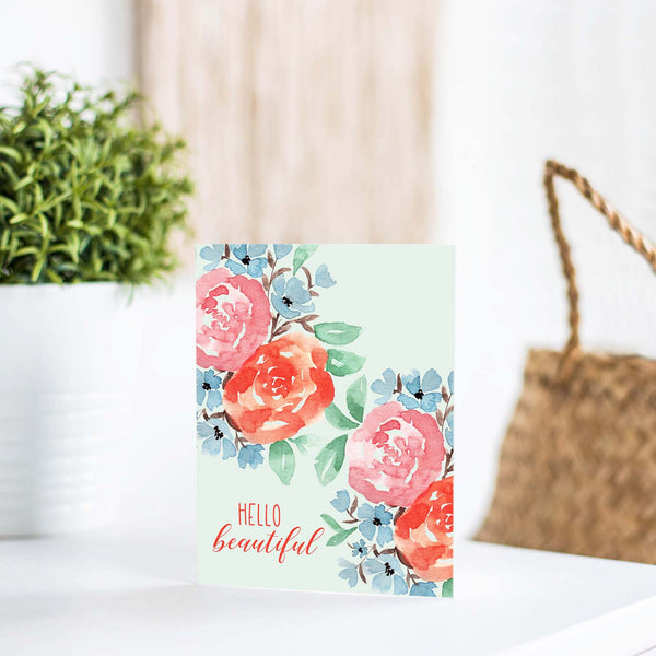 hello beautiful floral watercolor greeting card with coral, pink, blue flowers and green leaves with white A2 envelope shown on a white table with a plant and handbag
