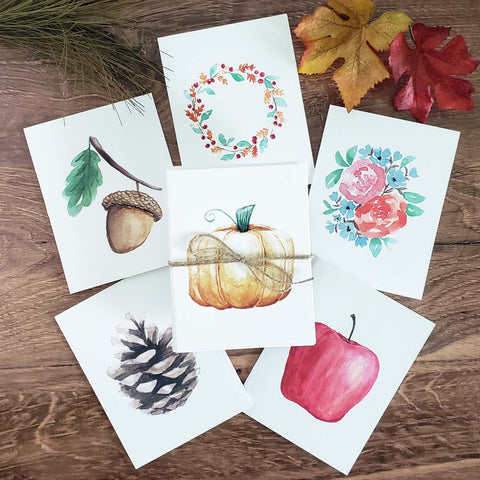 Set of 6 Watercolor painted notecards in fall autumn nature designs including an acorn with leaf, orange pumpkin, fall foliage wreath, red apple, fall floral bouquet and pinecone shown on a wooden table