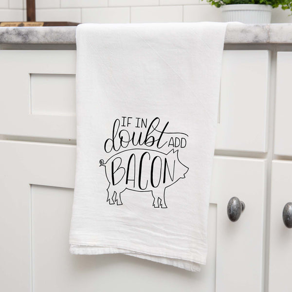 White floursack kitchen towel with black hand lettered illustrated design that says If in doubt add bacon with a outline of a pig shown folded and hanging from a countertop in a modern kitchen