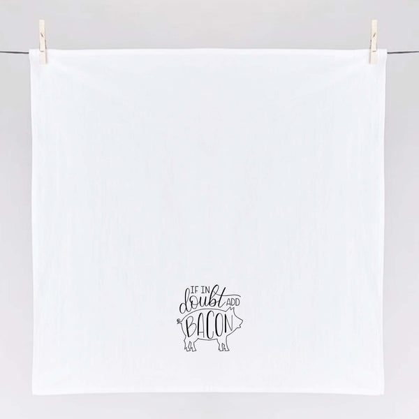 White floursack kitchen towel with black hand lettered illustrated design that says If in doubt add bacon with a outline of a pig shown unfolded and hanging from clothes pins