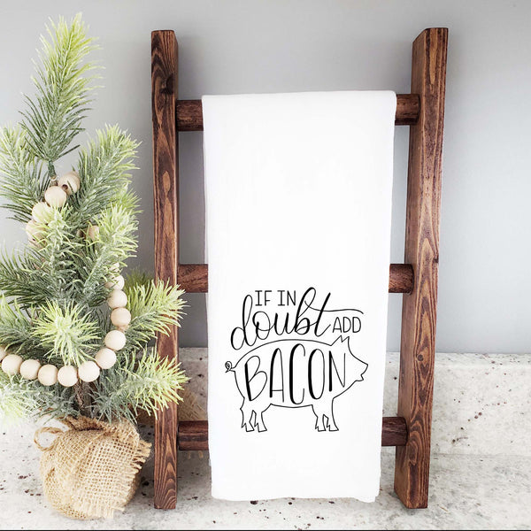 White floursack kitchen towel with black hand lettered illustrated design that says If in doubt add bacon with a outline of a pig shown folded and hanging from a wooden display ladder and a mini Christmas tree