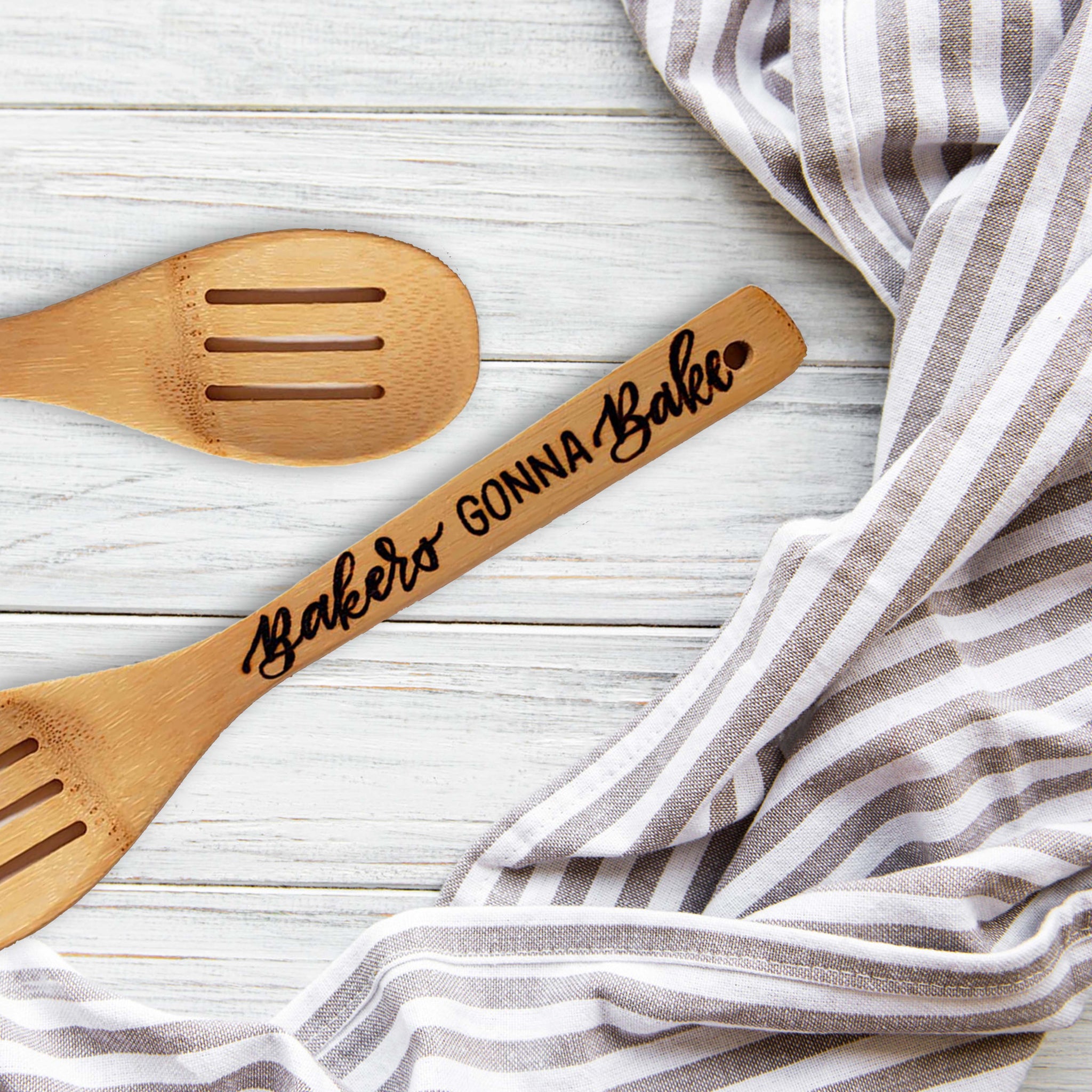 Bamboo wooden slotted spoon wood burned hand lettering that says Bakers Gonna Bake laying on a table with a towel