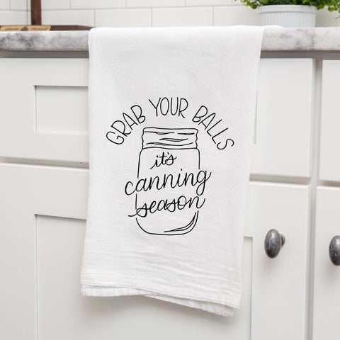 White floursack kitchen towel with black hand lettered illustrated design that says Grab your balls it's canning season with a mason jar doodle shown folded and hanging from a countertop in a modern kitchen
