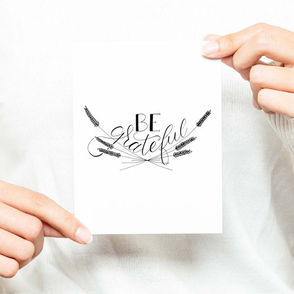 Be Grateful A2 Greeting Card, black hand lettered design with wheat illustrations on a folded white card shown with a woman in a white sweater holding card
