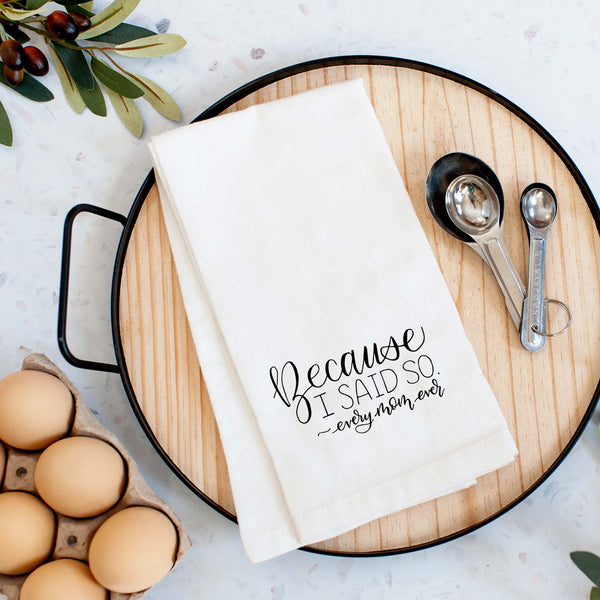 White floursack towel with black hand lettered illustrated design that says Because I said so - every mom ever shown folded on a serving tray with a set of measuring spoons and fresh eggs