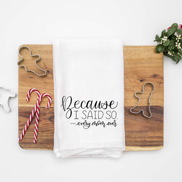 White floursack towel with black hand lettered illustrated design that says Because I said so - every mom ever shown folded on a wood cutting board with Christmas cookie cutters and candy canes
