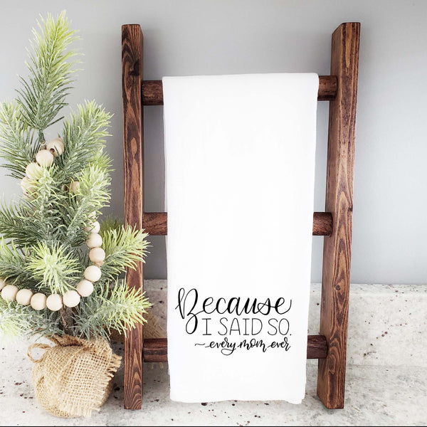 White floursack towel with black hand lettered illustrated design that says Because I said so - every mom ever shown folded and hanging from a wooden display ladder and a mini Christmas tree