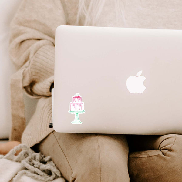 3" vinyl sticker of a watercolor painted 2-tiered white birthday cake with pink frosting and 3 cherries on top all sitting on a vintage scalloped mint colored cake stand shown adhered to a macbook laptop sitting on a woman's lap