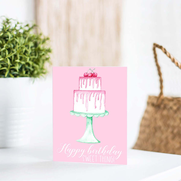 Happy birthday sweet thing watercolor birthday cake with pink frosting cherries and seafoam green cake stand birthday card shown on a white table with a plant and handbag