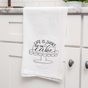 White floursack towel with black hand lettered illustrated design that says Life is short eat the cake with a cake on a stand doodle shown folded and hanging from a countertop in a modern kitchen