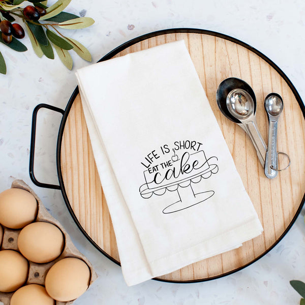 White floursack towel with black hand lettered illustrated design that says Life is short eat the cake with a cake on a stand doodle shown folded on a serving tray with a set of measuring spoons and fresh eggs