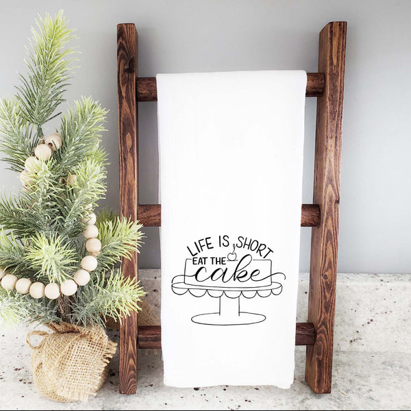 White floursack towel with black hand lettered illustrated design that says Life is short eat the cake with a cake on a stand doodle shown folded and hanging from a wooden display ladder and a mini Christmas tree