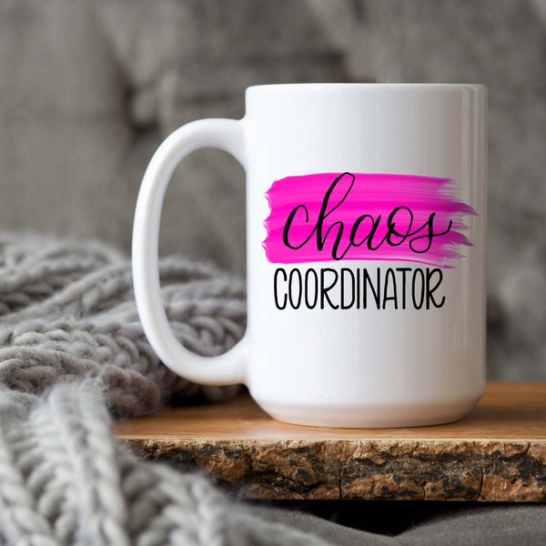 15oz white ceramic mug with hand lettered illustrated design that says chaos coordinator with a pink paint swash shown sitting on a wood tray with a grey knit blanket