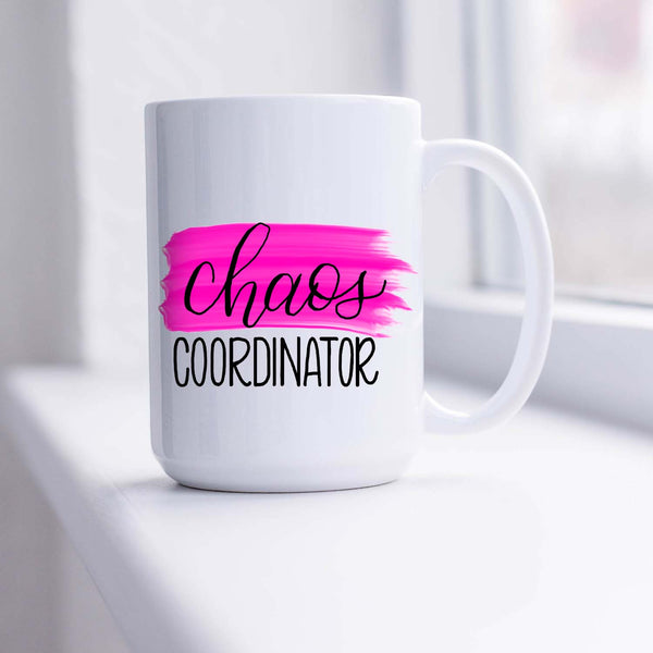 15oz white ceramic mug with hand lettered illustrated design that says chaos coordinator with a pink paint swash shown sitting in a sunny window