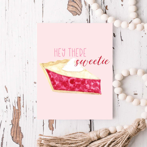 hey there sweetie watercolor greeting card with a watercolor slice of cherry pie topped with a dollop of whipped cream greeting card with white A2 envelope shown laying on a rustic white wooden table with white wooden bead garland