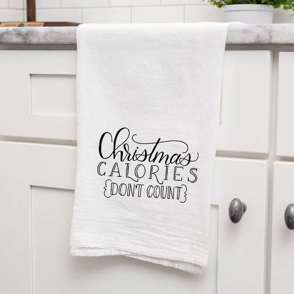 White floursack towel with black hand lettered illustrated design that says Christmas calories don't count shown folded and hanging from a countertop in a modern kitchen