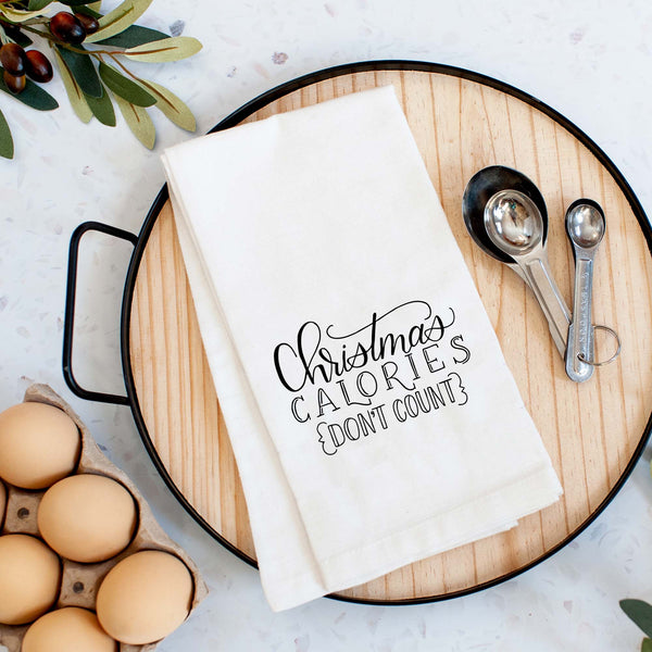 White floursack towel with black hand lettered illustrated design that says Christmas calories don't count shown folded on a serving tray with a set of measuring spoons and fresh eggs