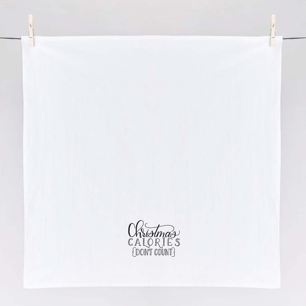White floursack towel with black hand lettered illustrated design that says Christmas calories don't count shown unfolded and hanging from clothes pins