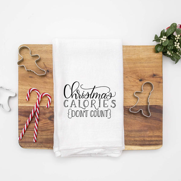 White floursack towel with black hand lettered illustrated design that says Christmas calories don't count shown folded on a wood cutting board with Christmas cookie cutters and candy canes