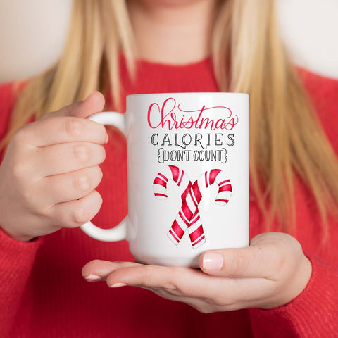 15oz white ceramic mug with watercolor candy canes and says christmas calories don't count in hand lettering shown with a woman in red sweater holding cup