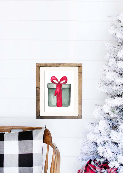 Watercolor painting of a green christmas package tied with a red bow shown in a wood frame hanging on a shiplap wall with a white flocked tree and rocking chair