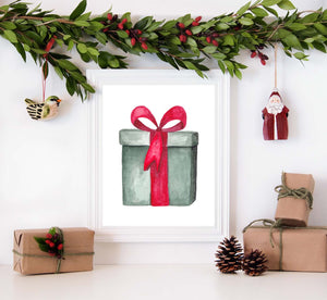 Watercolor painting of a green christmas package tied with a red bow shown in a white frame surrounded by christmas decorations
