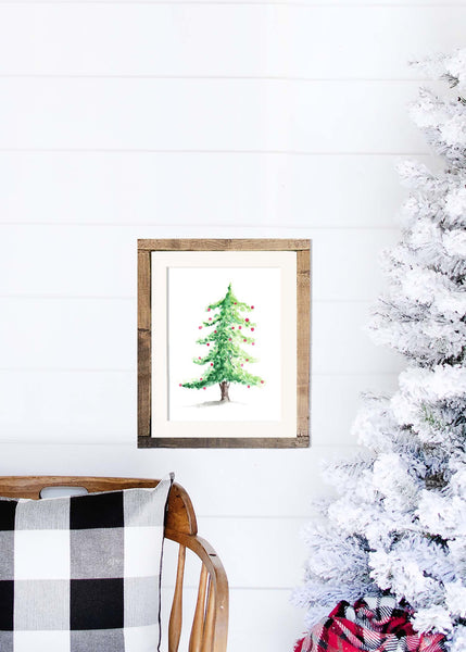 Watercolor woodland Christmas tree with red christmas bulbs shown in a wood frame hanging on a shiplap wall with a white flocked tree and rocking chair.