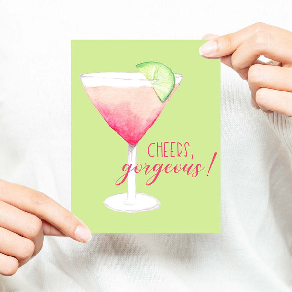 watercolor pink martini with lime in a cocktail glass friendship greeting card that says cheers gorgeous with a white A2 envelope shown with a woman in a white sweater holding card 