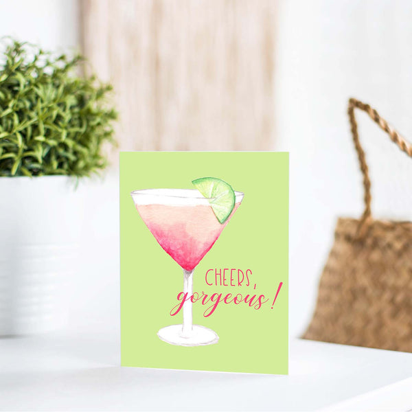 watercolor pink martini with lime in a cocktail glass friendship greeting card that says cheers gorgeous with a white A2 envelope shown standing on a white table with a white plant and handbag