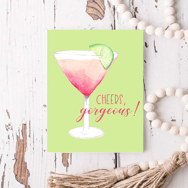 watercolor pink martini with lime in a cocktail glass friendship greeting card that says cheers gorgeous with a white A2 envelope shown laying on a rustic white wood table with white wood beaded garland