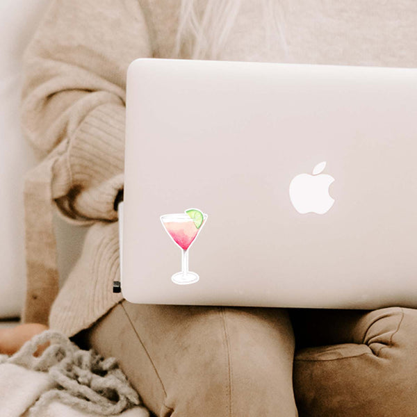 3" vinyl sticker of a watercolor martini glass with a pink cocktail and a slice of lime for garnish shown adhered to a MacBook sitting open on a woman's lap