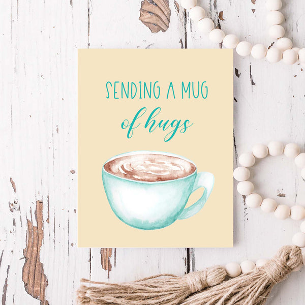 blue watercolor coffee mug filled with coffee friendship card that says sending a mug of hugs with a white A2 envelope shown laying on a rustic wooden table with white wooden bead garland