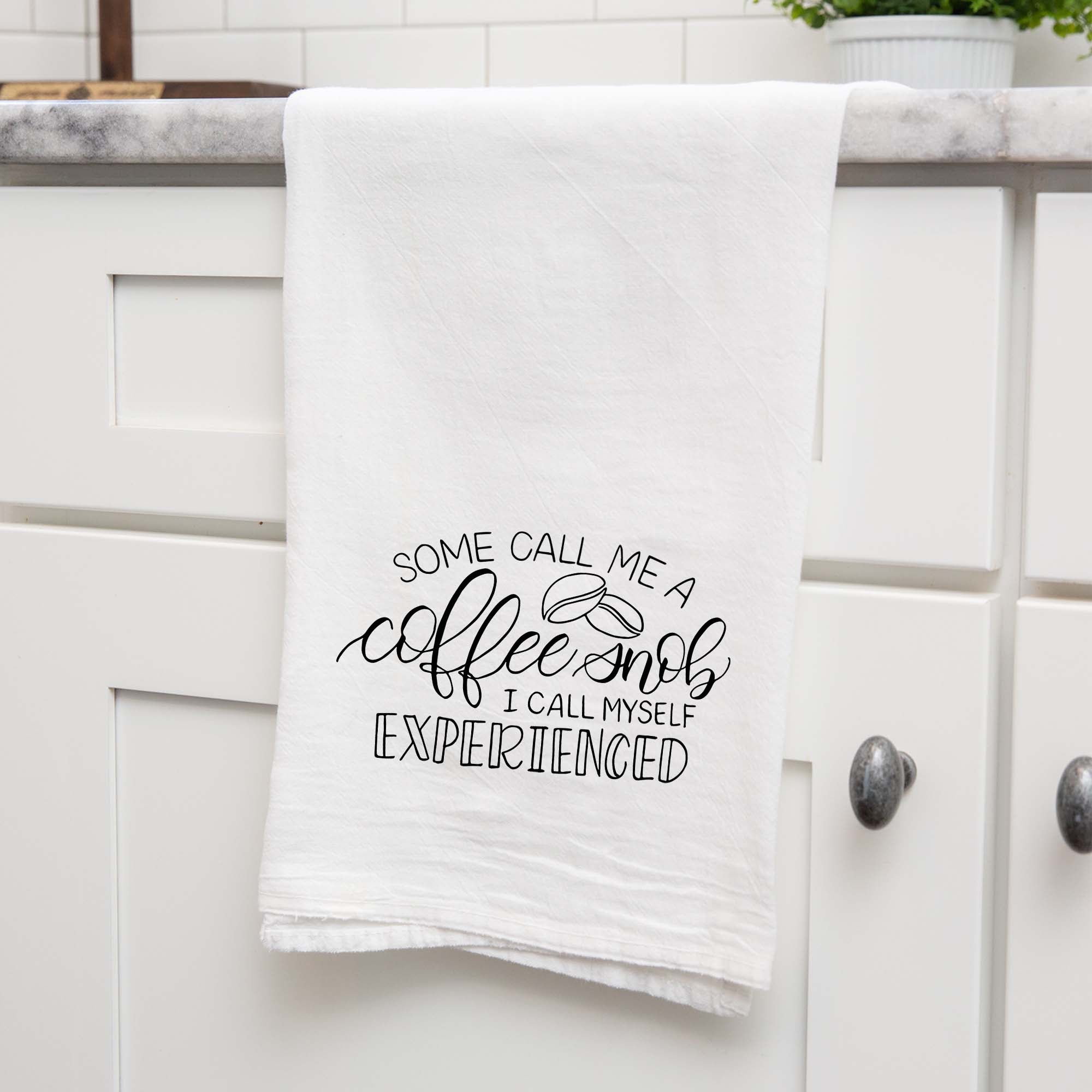 White floursack towel with black hand lettered illustrated design that says Some call me a coffee snob, I call myself experienced with coffee bean doodles shown folded and hanging from a countertop in a modern kitchen