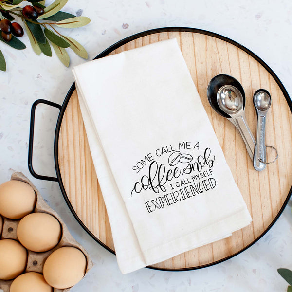 White floursack towel with black hand lettered illustrated design that says Some call me a coffee snob, I call myself experienced with coffee bean doodles shown folded on a serving tray with a set of measuring spoon and fresh eggs