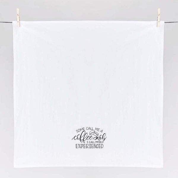 White floursack towel with black hand lettered illustrated design that says Some call me a coffee snob, I call myself experienced with coffee bean doodles shown unfolded and hanging from clothes pins