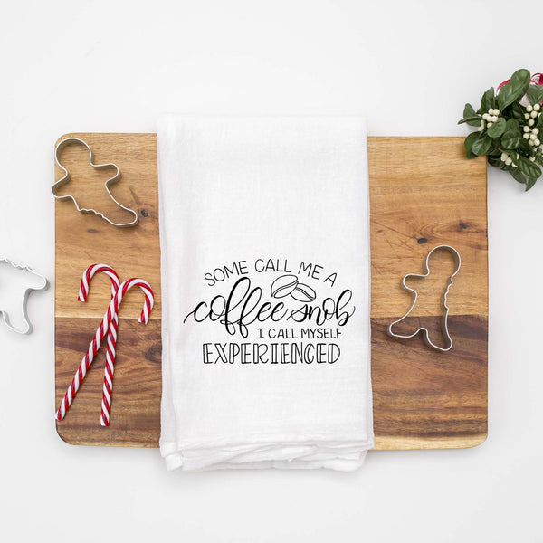 White floursack towel with black hand lettered illustrated design that says Some call me a coffee snob, I call myself experienced with coffee bean doodles shown folded on a wood cutting board with Christmas cookie cutters and candy canes