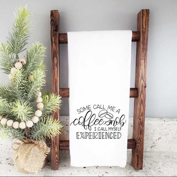 White floursack towel with black hand lettered illustrated design that says Some call me a coffee snob, I call myself experienced with coffee bean doodles shown folded and hanging from a wooden display ladder  and mini Christmas tree