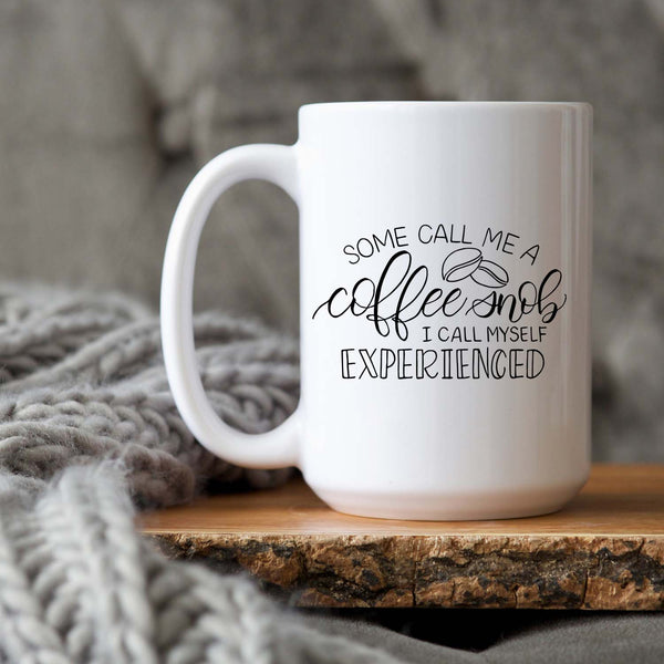 15oz white ceramic mug with hand lettered illustrated design that says some call me a coffee snob I call myself experienced shown sitting on a wood tray with a grey knit blanket