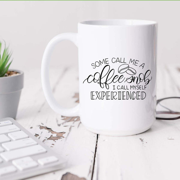 15oz white ceramic mug with hand lettered illustrated design that says some call me a coffee snob I call myself experienced shown on a white office desk