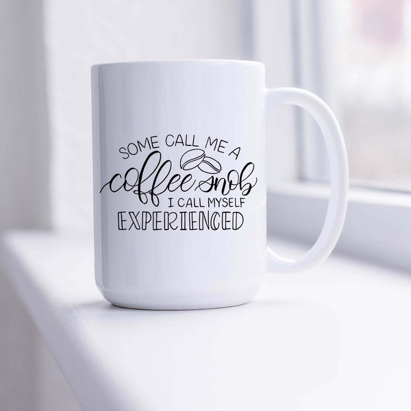 15oz white ceramic mug with hand lettered illustrated design that says some call me a coffee snob I call myself experienced shown in a sunny window