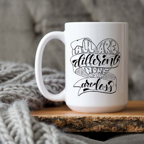 15oz white ceramic mug with hand lettered illustrated design that says all of us are different none of us are less shown on a wooden tray and grey knit blanket