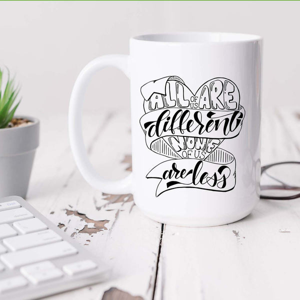 15oz white ceramic mug with hand lettered illustrated design that says all of us are different none of us are less sitting on a white desk with a plant, keyboard and glasses