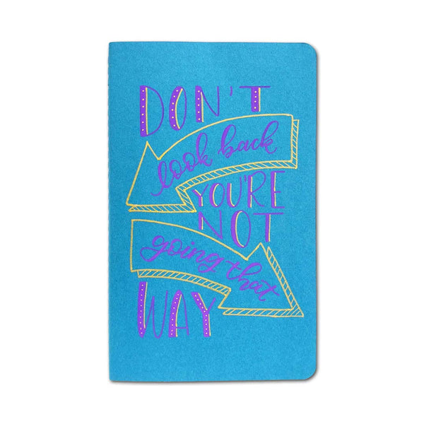 hand painted journal cover that says don't look back you're not going that way in block lettering and modern calligraphy with arrow doodles shown on the teal journal cover and purple and gold paint.