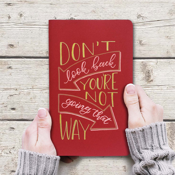 hand painted journal cover that says don't look back you're not going that way in block lettering and modern calligraphy with arrow doodles shown on the cranberry journal cover and gold and pink paint shown with woman's hand holding it